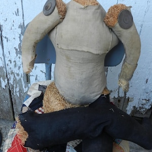 Puede incluir: Un torso de espantapájaros vintage hecho a mano, con tela de arpillera y detalles de paja. Las piernas negras están sobre un edredón de retazos. El conjunto evoca una estética rústica y campestre.