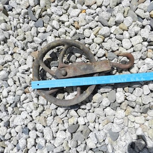 Puede incluir: Una polea de metal oxidado con un gancho, que mide aproximadamente 15 cm de diámetro, está sobre un lecho de pequeñas piedras grises.