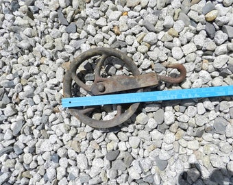 May include: A rusty metal pulley with a hook, measuring approximately 6 inches in diameter, is lying on a bed of small gray stones.