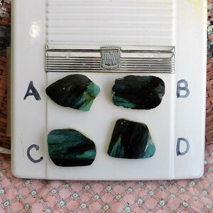以下が含まれることがあります： 白い表面にA、B、C、Dとラベル付けされた、緑と黒の虹色のガラスの破片4つ