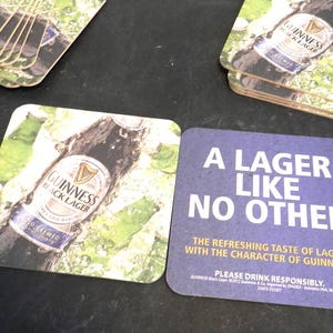 May include: Two square coasters featuring a bottle of Guinness Black Lager. One coaster has a photo of the bottle and the other has the text "A Lager Like No Other. The refreshing taste of lager with the character of Guinness."  Please drink responsibly.