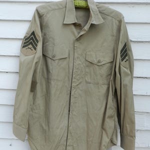 Könnte beinhalten: Ein militärisch anmutendes Hemd in Beige mit zwei Brusttaschen und Knopfverschlüssen. Das Hemd hat dunkelgrüne Winkel auf den Ärmeln, die den Rang anzeigen. Es hängt an einem Metallbügel vor einem weißen Holzgrund.