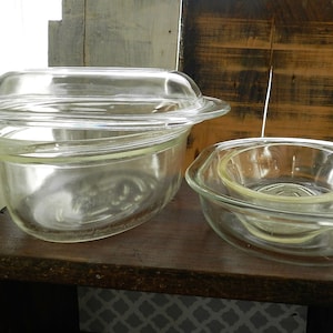 May include: Two clear glass baking dishes, one with a lid, on a wooden shelf. The dishes are oval and round in shape.