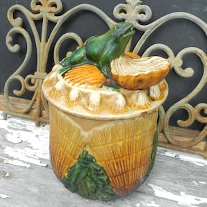 Könnte beinhalten: Eine Keramikdose mit einem grünen Froschdeckel. Die Dose ist mit einem gelben und braunen Blumendesign verziert. Der Frosch sitzt auf einer gelben und braunen Blume.