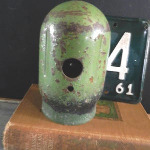 Puede incluir: Un objeto metálico verde vintage con una parte superior redondeada y un agujero circular. El objeto tiene un aspecto envejecido y desgastado con óxido visible. Una placa de matrícula verde con el número 4 y el número 61 está en el fondo. El objeto está sobre un libro.