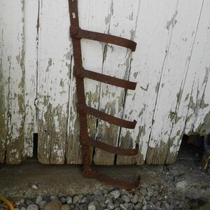 Puede incluir: Un objeto de metal oxidado con forma de escalera con cinco peldaños, apoyado contra una pared de madera blanca con pintura descascarada. El objeto descansa sobre un lecho de pequeñas piedras grises.