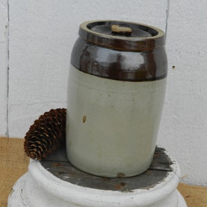 Puede incluir: Un tarro de cerámica vintage blanco y marrón glaseado con una tapa de madera. El tarro está sentado sobre un pedestal blanco.