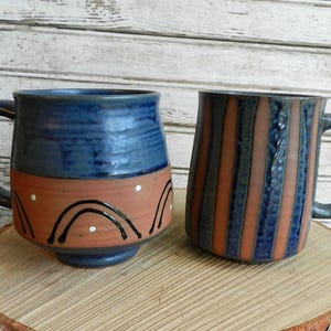 May include: Two handmade ceramic mugs with blue and orange glaze. One mug has a geometric pattern and the other has a more abstract design.