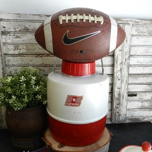 Könnte beinhalten: Ein weißer und roter Vintage-Kühlbehälter mit einem schwarzen und goldenen "Hawthorne"-Logo. Ein brauner und weißer Fußball liegt oben auf dem Kühlbehälter.