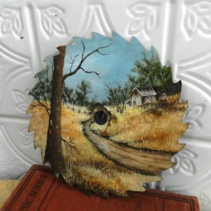 Puede incluir: Una hoja de sierra circular transformada en una pintura de paisaje rústico. La obra de arte representa un camino de tierra que conduce a una granja, con un árbol a la izquierda y un cielo azul. Los dientes de la hoja de sierra son visibles en el borde.