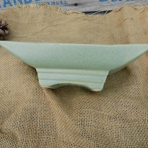 May include: A light green ceramic planter with a rectangular base and a wide, shallow bowl. The planter has a speckled glaze and a slightly textured surface.