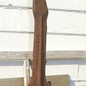 Puede incluir: Una herramienta de metal oxidado y desgastado, con un eje largo y rectangular y una parte superior plana y ensanchada. La base está unida a un bloque de madera. El texto "DEC 25 1900" es visible en el lateral. La herramienta parece ser antigua.