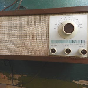 Może przedstawiać: Radio w stylu vintage z drewnianą ramą i tkaną osłoną głośnika. Biały panel przedni posiada pokrętło strojenia, regulację głośności, basów i tonów wysokich. Pokrętło jest brązowe, a tekst czarny.