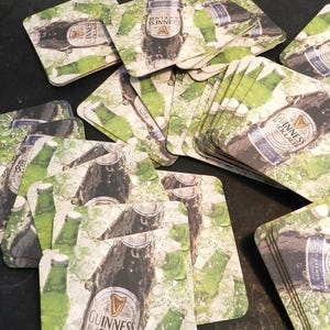 May include: A stack of square brown cardboard coasters with a green and white design featuring a bottle of Guinness Black Lager. The coasters have a water droplet effect.