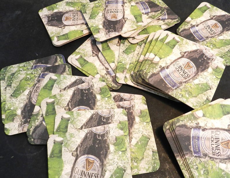 May include: A stack of square brown cardboard coasters with a green and white design featuring a bottle of Guinness Black Lager. The coasters have a water droplet effect.