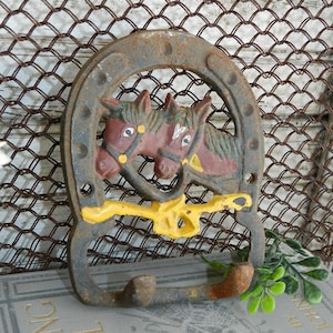 Puede incluir: Una herradura de hierro fundido oxidada con dos caballos pintados en rojo, amarillo y verde, con una cinta amarilla y dos ganchos en la parte inferior.