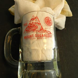 May include: A clear glass mug with a red and white design. The mug features a mountain scene with the text "Mount Sugarloaf South Deerfield 1974". A Rotary International logo is also visible on the mug.