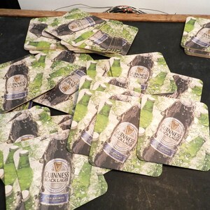 May include: A pile of square, brown cardboard coasters with a green, white, and black design. The coasters feature a bottle of Guinness Black Lager with the text "GUINNESS BLACK LAGER COLD BREWED" printed on the bottle.