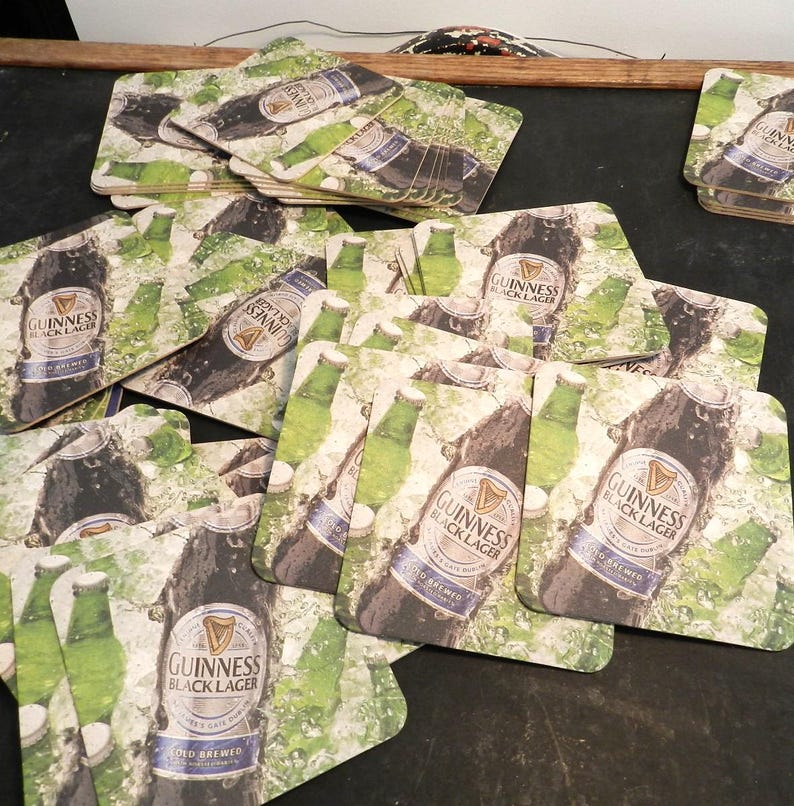 May include: A pile of square, brown cardboard coasters with a green, white, and black design. The coasters feature a bottle of Guinness Black Lager with the text "GUINNESS BLACK LAGER COLD BREWED" printed on the bottle.