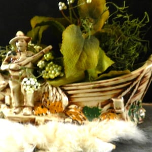 Puede incluir: Una maceta decorativa de cerámica en forma de barco con una figura tocando un instrumento de cuerda. La maceta es de color beige y marrón, con detalles en naranja y verde. Está llena de vegetación artificial y bayas blancas.