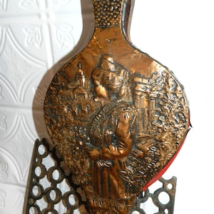 Puede incluir: Antiguo fuelle de cobre con un diseño en relieve detallado. El fuelle presenta un óvalo central con un relieve de una figura en un paisaje y un asa texturizada. Una pieza decorativa para una chimenea.