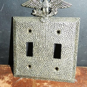 Puede incluir: Una cubierta de interruptor de luz doble de estilo vintage, en tono plateado, con una superficie texturizada. Un emblema de águila se encuentra en la parte superior de la cubierta. La cubierta tiene dos aberturas rectangulares y cuatro orificios para tornillos.