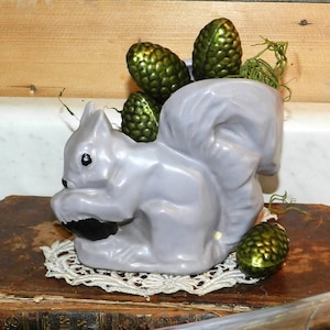 May include: A light grey ceramic squirrel-shaped planter with green decorative pinecones. The squirrel sits on a white lace doily, placed on a vintage book. The planter has a glossy finish.