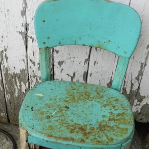 Puede incluir: Una silla de metal turquesa vintage con respaldo curvo y asiento redondo. La silla muestra signos de edad con óxido y desgaste. El fondo presenta tablas de madera blancas desgastadas.