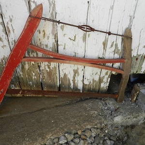 Puede incluir: Una sierra de madera roja con una hoja de metal y un tensor de alambre. La sierra está apoyada contra una pared de madera blanca.