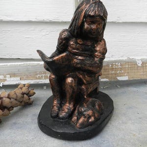 Puede incluir: Una estatua de bronce de una niña sentada leyendo un libro con un perro a sus pies. La estatua está sobre una base negra.