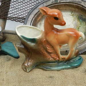 Puede incluir: Un macetero de cerámica con forma de tronco de árbol con una figurita de ciervo marrón de pie encima. El ciervo es marrón con detalles blancos y tiene una base verde.