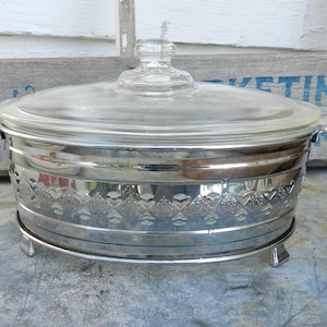 Peut inclure: Un plat à casserole argenté vintage avec un couvercle et des poignées en verre transparent. Le plat présente un motif gravé décoratif au milieu et repose sur de petits pieds. Une pièce classique pour servir et recevoir.