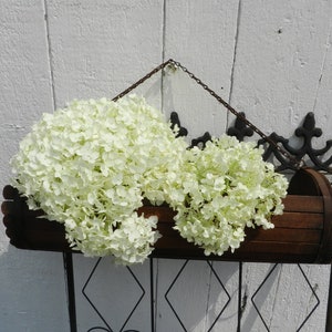 May include: Two white hydrangea flower arrangements in a wooden planter hanging from a black metal gate. The gate is against a white wooden wall.