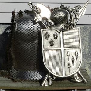 Puede incluir: Un conjunto de armadura de caballero de metal plateado con un escudo, un casco y una espada. El escudo tiene un escudo de armas con flores de lis y cruces.