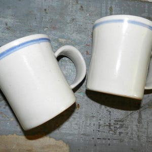 May include: Two white ceramic mugs with a blue stripe near the rim. Each mug has a curved handle and a slightly tapered shape. The mugs are displayed on a weathered, gray wooden surface.