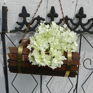 May include: A white flower arrangement hangs from a black metal gate. The flowers are in a wooden planter with gold metal bands.