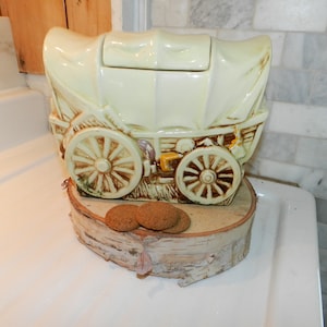 May include: A yellow ceramic cookie jar shaped like a covered wagon with brown wheels and a wooden base. There are three chocolate chip cookies on the base.