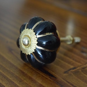 May include: Black and gold ceramic cabinet knob with a decorative, fluted design. The knob has a gold centre with a raised, circular design.