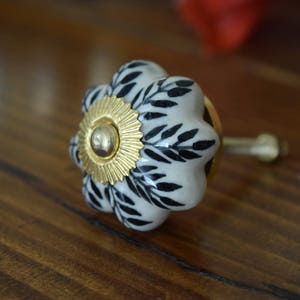 May include: White ceramic cabinet knob with a black floral design and a gold center. The knob has a brass screw.