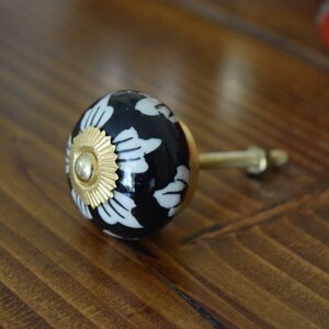 May include: A black and white ceramic knob with a gold accent and a floral design. The knob is attached to a metal stem.