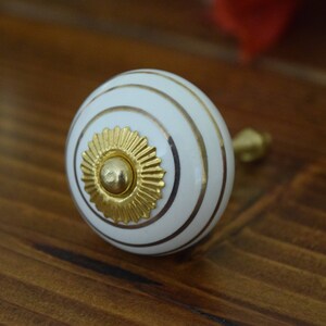 May include: White ceramic knob with gold trim and a gold center piece. The knob has a circular shape and is designed for use on cabinets or drawers.