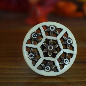 May include: A white round decorative knob with a carved floral design in brown. The knob has a geometric pattern with a central flower.