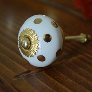 May include: White ceramic knob with gold polka dots and a gold decorative centre. The knob has a brass screw.