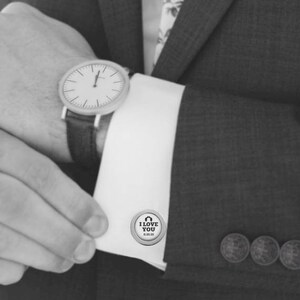 May include: Silver cufflinks with a circular design featuring the text "I love you" and a heart symbol. The cufflinks are shown on a white shirt cuff.