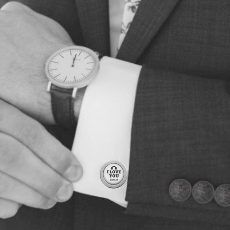 May include: Silver cufflinks with a circular design featuring the text "I love you" and a heart symbol. The cufflinks are shown on a white shirt cuff.
