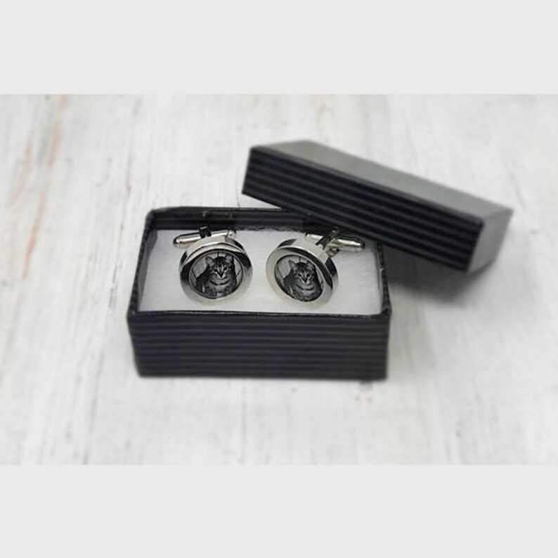 May include: A pair of silver cufflinks with a black and white photo of a cat inside each cufflink. The cufflinks are in a black box with a striped pattern.