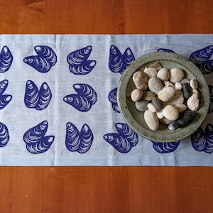 May include: A light grey table runner featuring a repeating pattern of dark blue mussel shell illustrations. A round, textured, sage green ceramic bowl filled with a collection of smooth, varied-coloured river stones sits on the fabric surface.