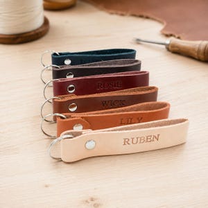May include: A stack of six leather keychains in varying shades of blue, dark brown, burgundy, medium brown, tan, and natural beige. Each keychain features a silver metal snap button, a silver split ring, and a name embossed on the leather surface.