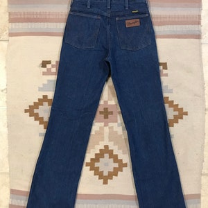 May include: A pair of dark blue denim jeans with a brown leather Wrangler label on the back pocket.