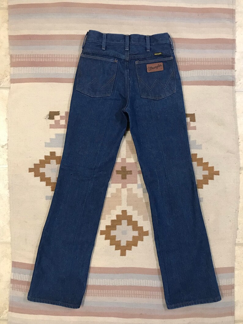 May include: A pair of dark blue denim jeans with a brown leather Wrangler label on the back pocket.